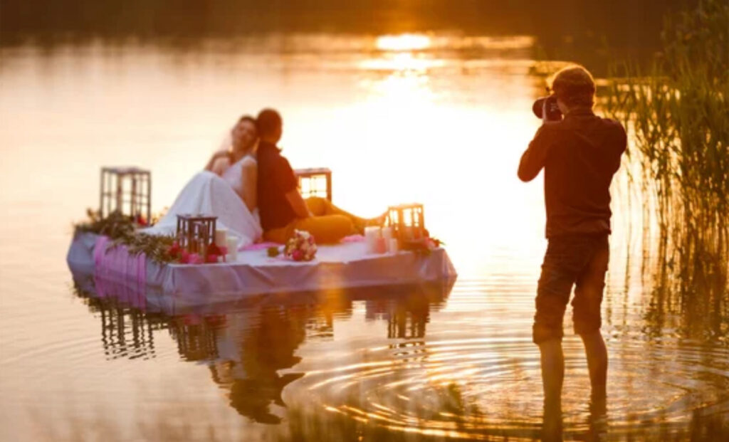 Wedding photographer in action, taking a picture of the bride and groom sitting on the raft. Summer, sunset
